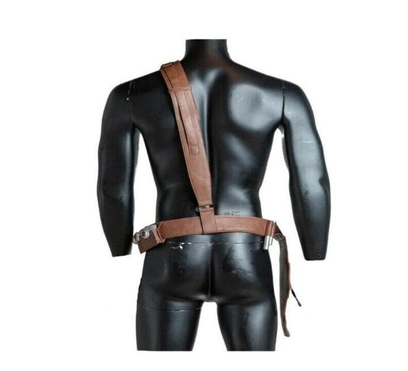 May include: A brown leather-like utility belt and shoulder strap harness worn across the torso. The set features a horizontal waist belt with attached metallic hardware components and a single vertical strap extending over the right shoulder.