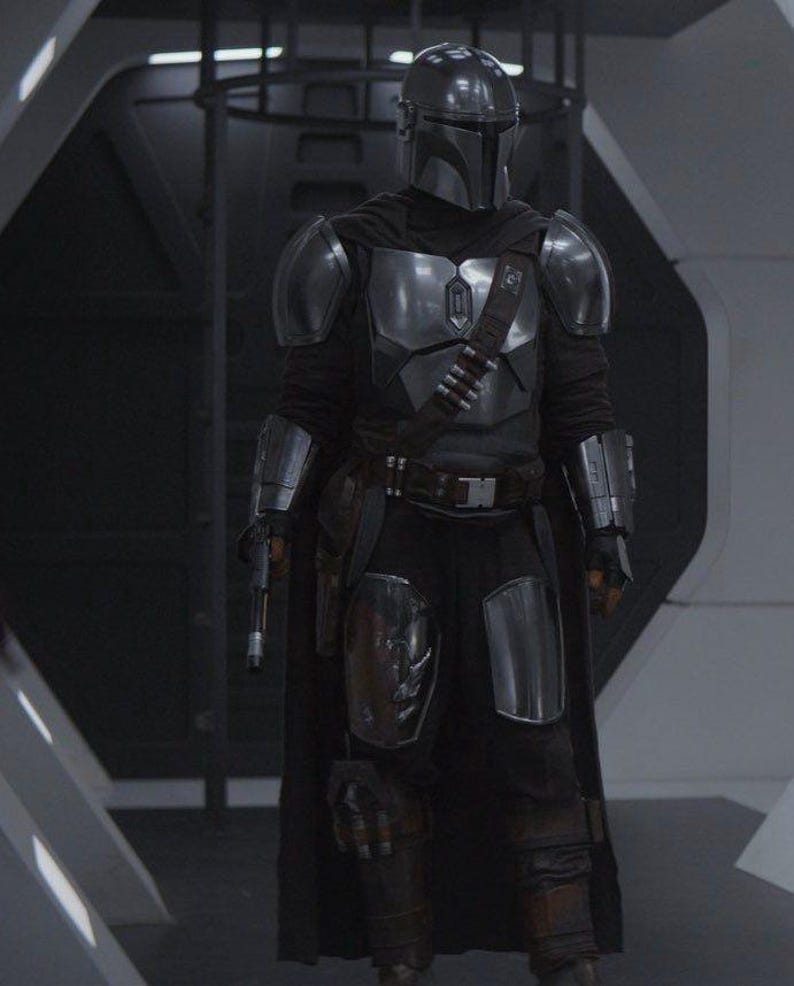 May include: A full-body shot of a character in a silver and black Mandalorian armor suit. The helmet is a polished silver, and the armor plates are detailed. The character is holding a blaster pistol. The background is a futuristic interior.