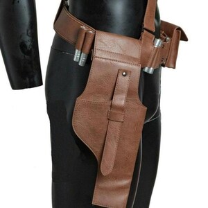 May include: A brown textured utility belt featuring a holster with a vertical strap and silver-toned cylindrical accents attached to the side. The belt includes a side pouch and is worn around the waist, constructed from a durable material.