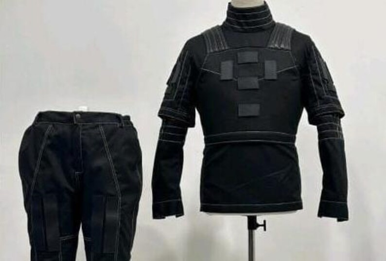 May include: A black tactical-style outfit consisting of a long-sleeved top with a high collar, shoulder padding, and rectangular chest patches, paired with matching trousers featuring reinforced leg panels and prominent white contrast stitching.