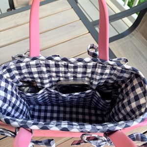 May include: A pink tote bag with a black and white gingham fabric lining. The interior features three open pockets containing a smartphone and two pairs of eyeglasses. The bag has two solid pink handles and small fabric ties on the sides.