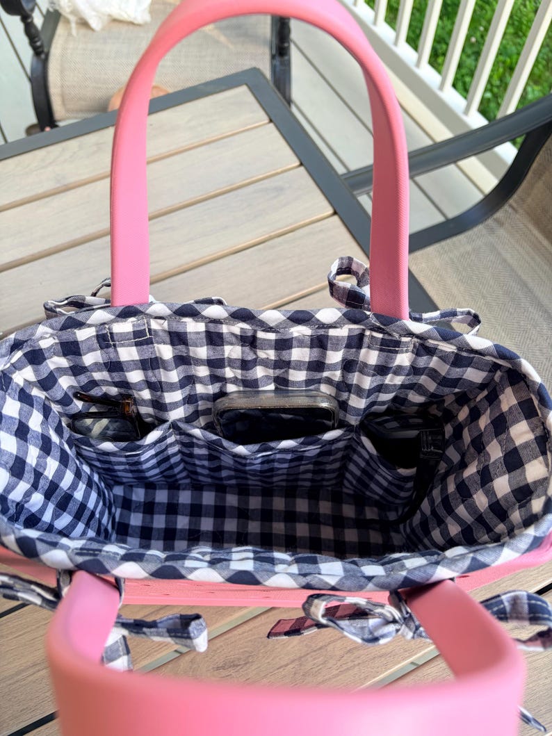 May include: A pink tote bag with a black and white gingham fabric lining. The interior features three open pockets containing a smartphone and two pairs of eyeglasses. The bag has two solid pink handles and small fabric ties on the sides.