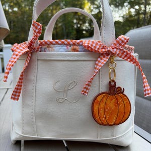 May include: A cream-coloured canvas tote bag featuring a stitched letter L on the front. The bag is accented with orange and white gingham ribbon bows tied at the top handles and includes a hanging orange pumpkin charm with a gold-tone clasp.