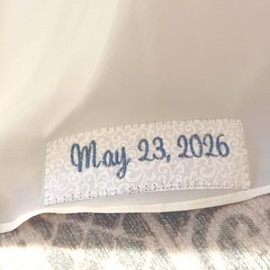 May include: A rectangular label with the text "May 23, 2026" in blue script. The label has a white background with a subtle floral pattern and is stitched around the edges.