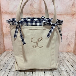 May include: A cream-coloured tote bag with a navy and white gingham lining and trim. The bag features a stitched "L" monogram in a light brown thread. It has two handles and is tied with gingham bows.