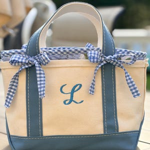 Può includere: Una borsa tote in tela color crema con una lettera L ricamata in blu sul davanti. La borsa presenta manici blu, una base blu e un bordo in tessuto a quadretti vichy blu e bianco lungo il bordo superiore, con due piccoli fiocchi sul davanti.