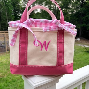 May include: A small canvas tote bag with a cream-colored body and dark pink trim. The bag features two pink handles, a pink embroidered letter W on the front, and a pink and white gingham fabric lining that ties into bows at the top sides.