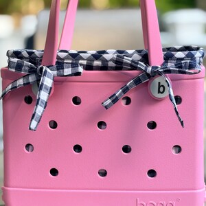 May include: A pink tote bag with a perforated exterior and two handles. The bag features a navy blue and white gingham fabric liner that extends over the top edge and is tied with matching gingham ribbons on each side. The word bogg is embossed.