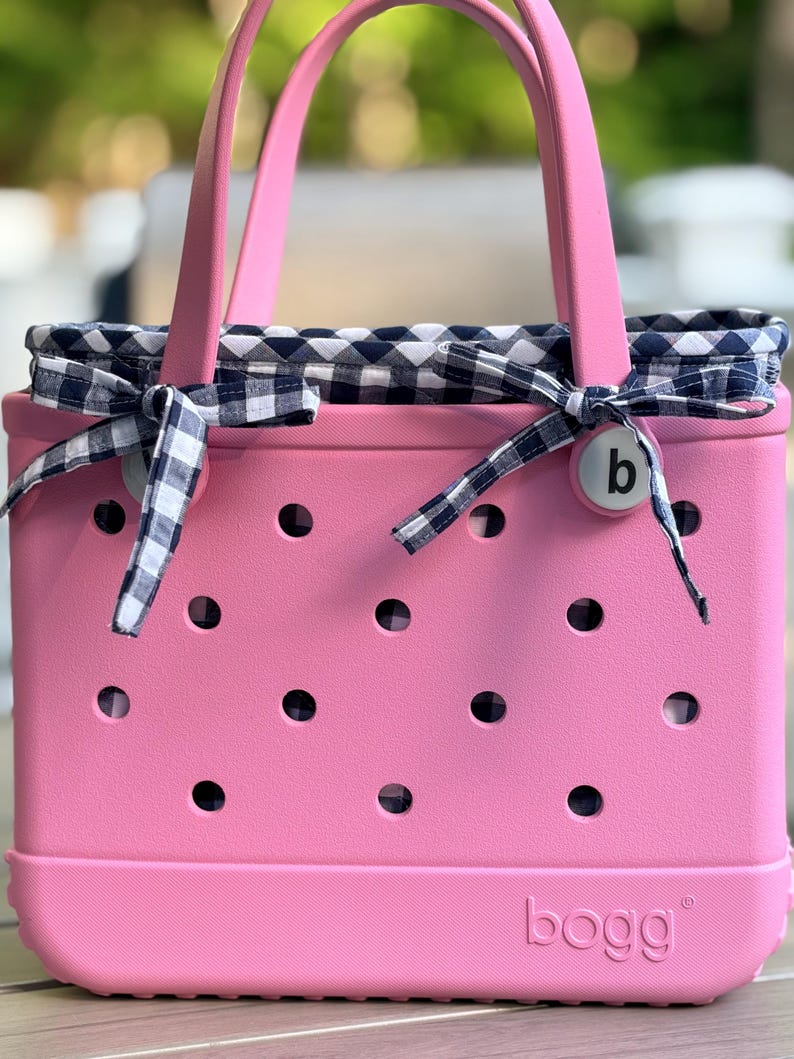 May include: A pink tote bag with a perforated exterior and two handles. The bag features a navy blue and white gingham fabric liner that extends over the top edge and is tied with matching gingham ribbons on each side. The word bogg is embossed.