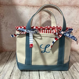 Könnte beinhalten: Kleine Canvas-Tragetasche mit cremefarbenem Korpus, blauen Akzenten und Griffen. Die Tasche hat ein rot-weißes Vichy-Futter und ist mit dekorativen Schleifen verziert. Auf der Vorderseite ist ein gestickter blauer Buchstabe "L".