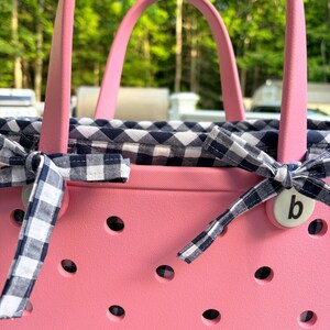 May include: A pink rubber tote bag featuring a perforated exterior and two sturdy handles. The interior is lined with a navy blue and white gingham fabric, which is tied at the sides with matching gingham ribbons and a small white button with a letter b.