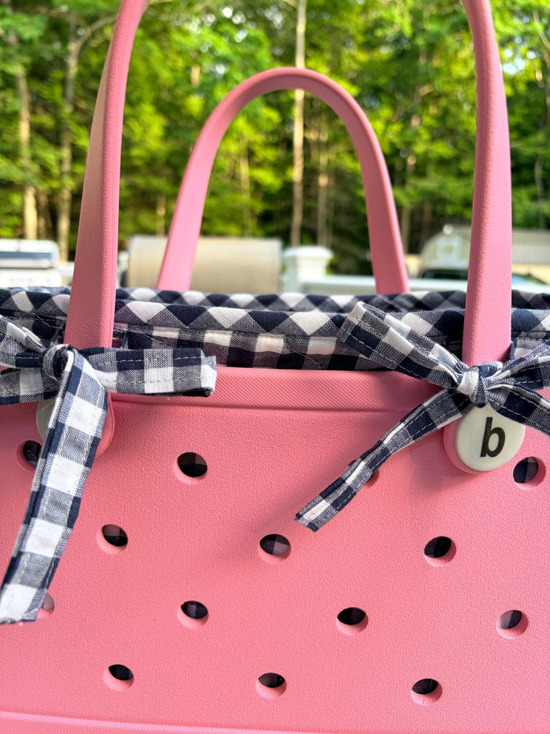 May include: A pink rubber tote bag featuring a perforated exterior and two sturdy handles. The interior is lined with a navy blue and white gingham fabric, which is tied at the sides with matching gingham ribbons and a small white button with a letter b.