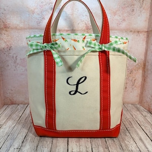 May include: A cream-coloured tote bag with red trim, handles, and base. It features a green and white gingham trim and a carrot-patterned lining. A navy blue script "L" is embroidered on the front. The bag has a classic, sturdy design.