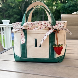 May include: A cream-coloured canvas tote bag with dark green trim and handles. The front features a dark green embroidered letter L. A floral fabric lining with a strawberry print extends from the top, tied with bows. A strawberry charm hangs.