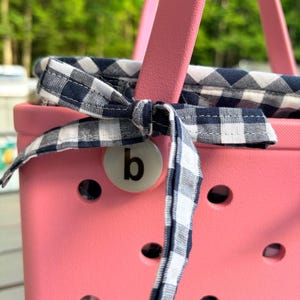May include: A bright pink rubber tote bag with a perforated surface and a pink handle. A navy blue and white gingham fabric liner is tied to the handle with a matching fabric bow. A circular white charm featuring the lowercase letter b is attached.