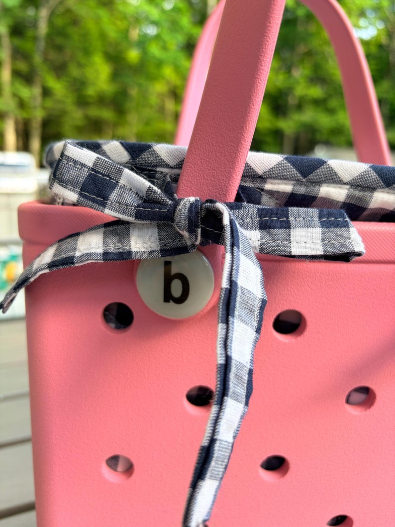 May include: A bright pink rubber tote bag with a perforated surface and a pink handle. A navy blue and white gingham fabric liner is tied to the handle with a matching fabric bow. A circular white charm featuring the lowercase letter b is attached.