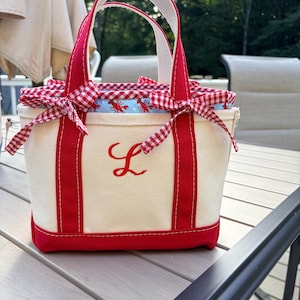 Puede incluir: Un bolso de lona color crema con ribetes y asas rojas. La parte frontal presenta una letra L bordada en rojo. La abertura superior está asegurada con una inserción de tela de cuadros rojos y blancos atada con lazos, con un estampado de langostas.