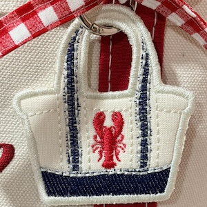 May include: A white canvas tote bag charm with a red lobster design. The charm has navy blue accents and a silver metal ring for attachment. A red and white gingham ribbon and a red stripe are visible in the background.