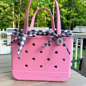 May include: A pink tote bag with a perforated body and two handles. The bag features a navy and white gingham fabric liner tied with bows at the top sides. A circular white button with the lowercase letter b is attached to the front right side.