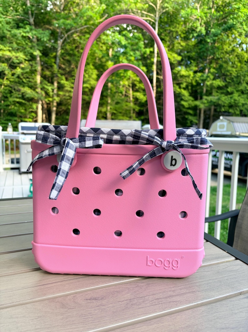 May include: A pink tote bag with a perforated body and two handles. The bag features a navy and white gingham fabric liner tied with bows at the top sides. A circular white button with the lowercase letter b is attached to the front right side.