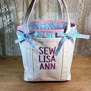 May include: A cream-coloured tote bag with a floral interior and accents. The bag features a pink trim, blue bows, and the embroidered text "SEW LISA ANN" in purple. It has two cream-coloured handles.
