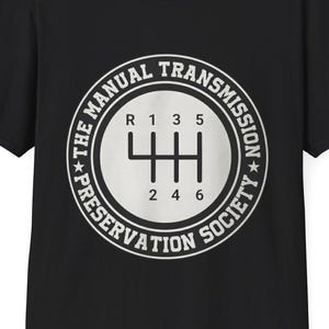May include: Black t-shirt with a white graphic of a manual transmission gear shift pattern. The graphic is encircled by the text "THE MANUAL TRANSMISSION PRESERVATION SOCIETY". The shirt has a crew neck and short sleeves.