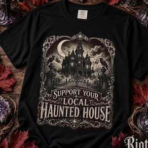 Puede incluir: Una camiseta negra con un estampado gráfico de una casa encantada de estilo victoriano, cuervos y una luna creciente dentro de un marco ornamentado. El texto dice "SUPPORT YOUR LOCAL HAUNTED HOUSE". Etiqueta de marca Comfort Colors en el cuello.