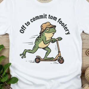Peut inclure: Un t-shirt blanc à manches courtes avec une illustration d'une grenouille verte portant un chapeau bob beige et montant une trottinette. Le texte au-dessus de la grenouille indique "Off to commit tom foolery" en noir. Étiquette de marque au col.