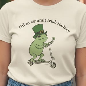 May include: A cream-coloured t-shirt with a green frog on a scooter, wearing a green top hat and holding a four-leaf clover. The text above the frog says, "Off to commit Irish foolery."