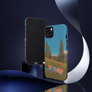 May include: Two phone cases with a landscape design. One case is black, and the other is light blue with a picnic scene. The picnic scene includes a blue blanket, a red and white checkered blanket, a picnic basket, and trees.