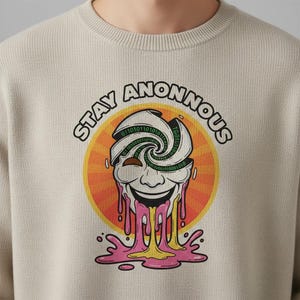 Pode incluir: Camisola bege de gola redonda com um gráfico de um rosto sorridente com código binário e as palavras "STAY ANONNOUS". O rosto tem detalhes gotejantes rosa e amarelo, contra um fundo de sol laranja e amarelo.