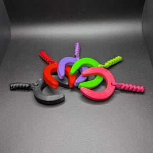 May include: A collection of colorful, C-shaped objects with braided cord handles. The objects are in various colors, including red, purple, green, pink, and black. The background is a dark gray surface.