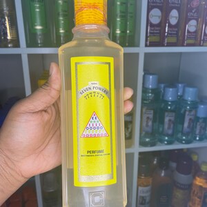 Peut inclure: Une bouteille de parfum transparente avec une étiquette jaune portant l'inscription "SEVEN POWERS" et une série de symboles. La bouteille a un bouchon jaune et est tenue dans une main. D'autres bouteilles sont visibles en arrière-plan.
