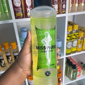 May include: A clear bottle of "Miss Paris Perfume" with a black cap, green label, and a white silhouette of a woman. The bottle contains a yellow liquid and is labeled 1000ml. Other bottles are visible on shelves in the background.