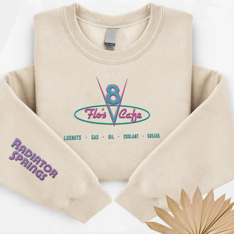 Puede incluir: Sudadera color crema con el texto bordado "Flo's Cafe" en un dise&ntilde;o turquesa y rosa. La manga presenta el texto "Radiator Springs" en una combinaci&oacute;n de colores similar. El dise&ntilde;o incluye las palabras "LUGNUTS GAS OIL COOLANT GREASE".