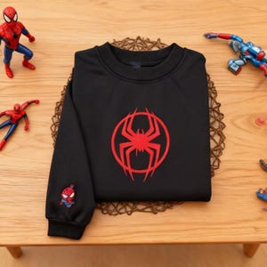 Pu&ograve; includere: Felpa nera con un logo di ragno rosso sul davanti e una grafica di Spider-Man sulla manica. La felpa &egrave; su un tavolo di legno, con action figure di Spider-Man.