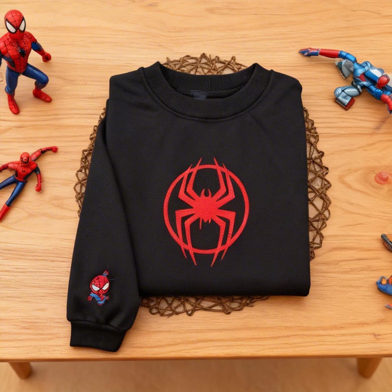 Pu&ograve; includere: Felpa nera con un logo di ragno rosso sul davanti e una grafica di Spider-Man sulla manica. La felpa &egrave; su un tavolo di legno, con action figure di Spider-Man.