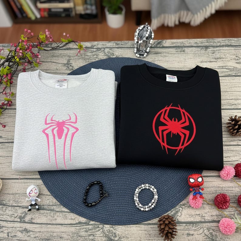 Pu&ograve; includere: Due felpe, una grigio chiaro e una nera, ciascuna con un'emblema di ragno. La felpa grigia ha un design di ragno rosa, mentre quella nera ha un design di ragno rosso all'interno di un cerchio rosso.