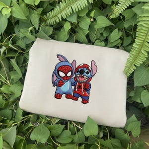 May include: Off-white tote bag featuring a cartoon illustration of two characters dressed as Spider-Man. One character is in a blue costume with a Spider-Man mask, and the other is in a red and blue hoodie. The bag is surrounded by green leaves.