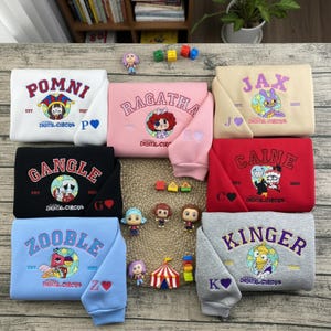 May include: A collection of folded sweatshirts in various colors, each featuring a unique design and character name. The sweatshirts display the text "POMNI", "RAGATHA", "JAX", "GANGLE", "CAINE", "ZOOBLE", and "KINGER", along with character illustrations and the words "DIGITAL CIRCUS".