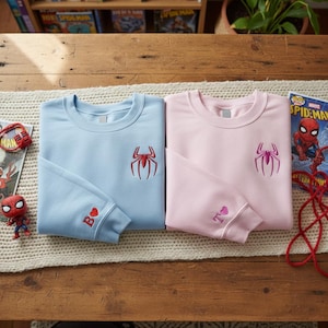 May include: Two crewneck sweatshirts, one light blue and one pink, each with a spider emblem embroidered on the chest. The light blue sweatshirt has a "B" and heart on the sleeve, and the pink one has a "T" and heart.