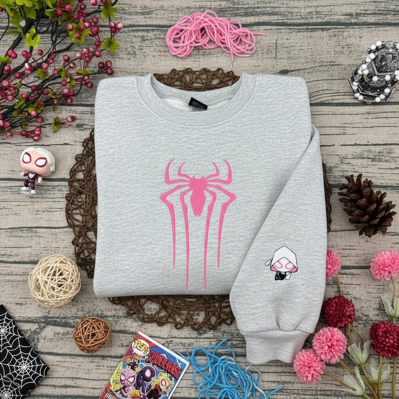 Pu&ograve; includere: Felpa grigio chiaro con un grande emblema di ragno rosa sul davanti e una grafica di Gwen Stacy sulla manica. La felpa ha girocollo e polsini e orlo a coste. Un fumetto &egrave; visibile.