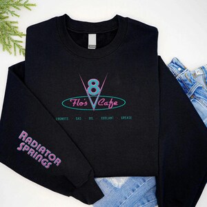 Puede incluir: Sudadera negra con el logo "Flo's Cafe" en verde azulado y rosa. La manga muestra "Radiator Springs" en rosa. Debajo del logo del caf&eacute; est&aacute;n las palabras "Lugnuts - Gas - Oil - Coolant - Grease".