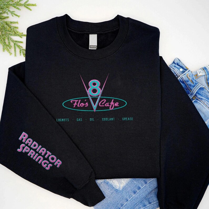 Puede incluir: Sudadera negra con el logo "Flo's Cafe" en verde azulado y rosa. La manga muestra "Radiator Springs" en rosa. Debajo del logo del caf&eacute; est&aacute;n las palabras "Lugnuts - Gas - Oil - Coolant - Grease".