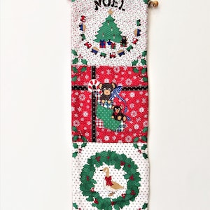 Pode incluir: Uma meia de Natal vermelha, branca e verde com um design acolchoado. A meia apresenta uma variedade de imagens festivas, incluindo uma árvore de Natal, ursos, uma coroa e um pato. A meia é decorada com flocos de neve e azevinho. A meia tem a palavra "NOEL" bordada.