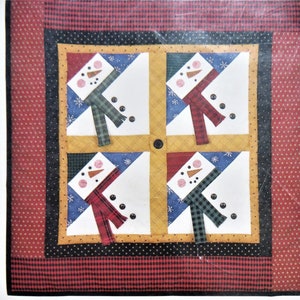 May include: A quilt pattern featuring four snowmen with different coloured scarves and hats. The quilt is made up of squares of red, white, blue, black and yellow fabric. The pattern is surrounded by a red and black plaid border. The text "EASY PROJECT!" is printed in black on a white background.