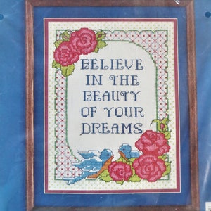May include: Counted cross stitch pattern featuring a blue background with a white frame. The pattern includes a floral design with red roses and blue birds. The text "Believe in the beauty of your dreams" is stitched in the center of the pattern. The finished size is 9 inches by 12 inches.