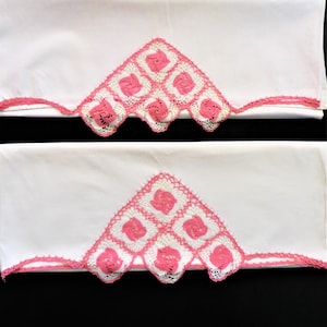 May include: Two white pillowcases with pink crocheted lace trim. The lace features a repeating pattern of squares with pink flowers in the centre.