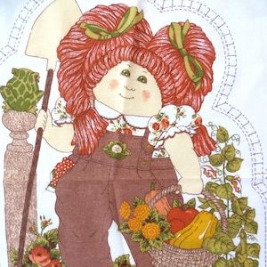 May include: A red-haired doll with a green bow in her hair is holding a shovel and a basket of vegetables. She is wearing a brown overall and a white shirt with a floral pattern. The doll is standing in a garden with green leaves and flowers. The text "Leave Open for Stuffing" is printed on the bottom of the image.
