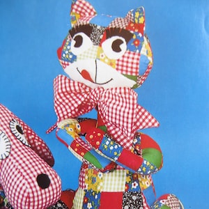 Può includere: Un giocattolo di gatto in patchwork con un fiocco a quadri rossi e bianchi. Il gatto è fatto di vari tessuti colorati, tra cui rosso, blu, verde, giallo e bianco. Il gatto ha grandi occhi neri e una lingua rosa che sporge.